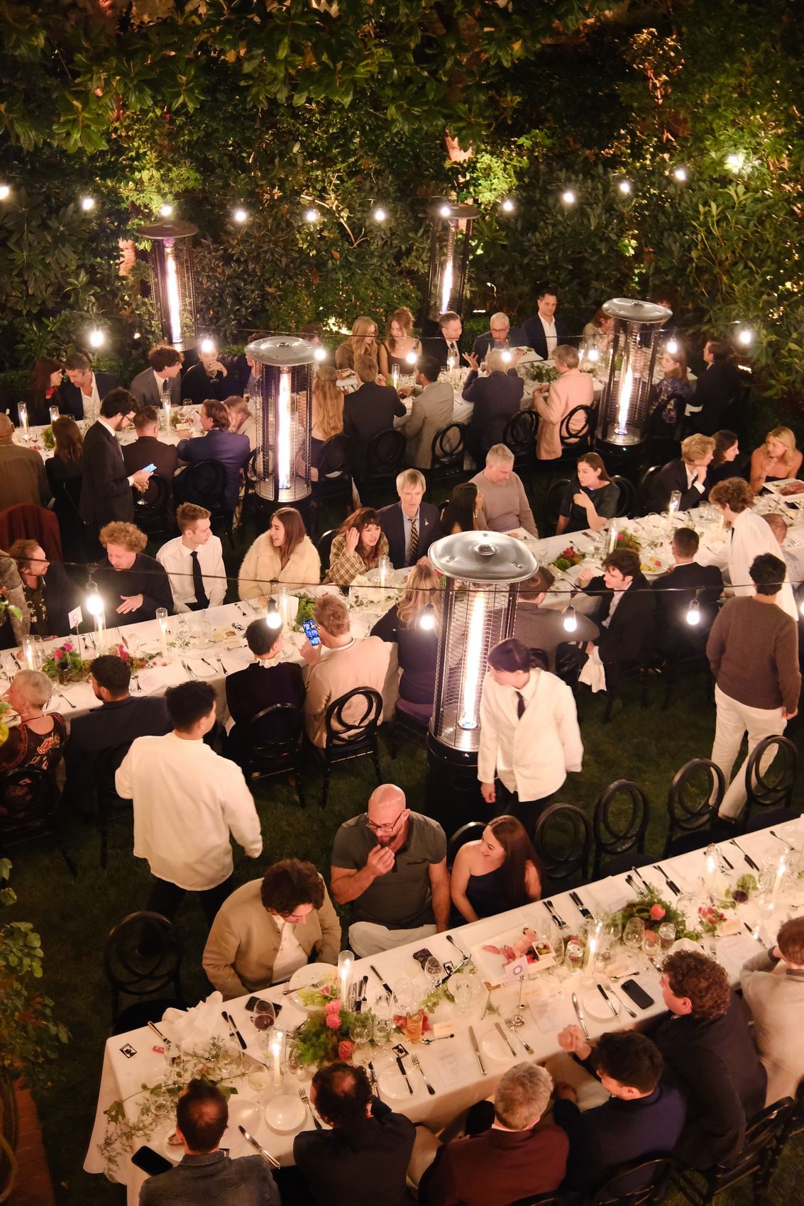 An overhead candlelit garden dinner with string lights and heaters