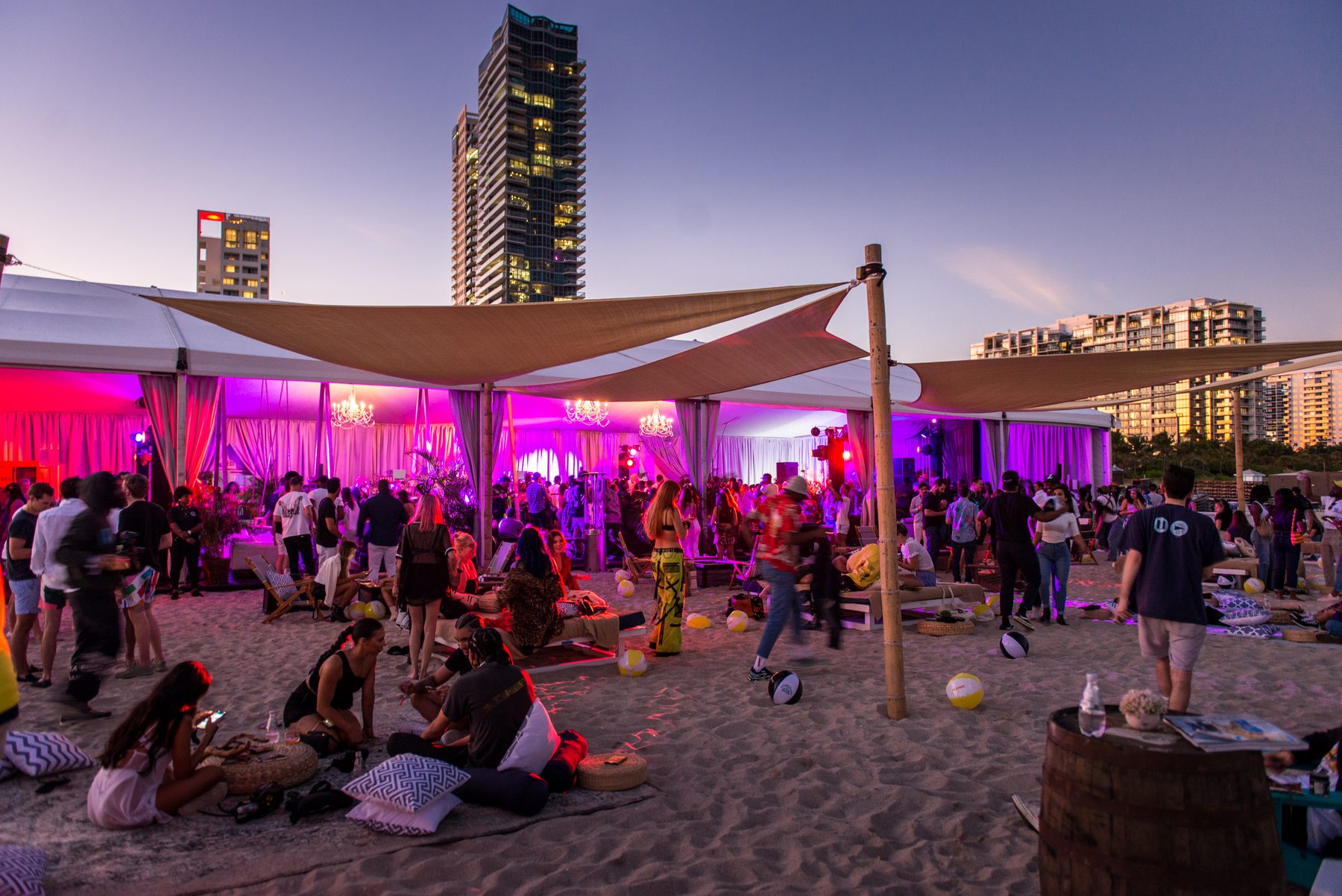 A beachfront party tent glowing pink at dusk with a city skyline behind