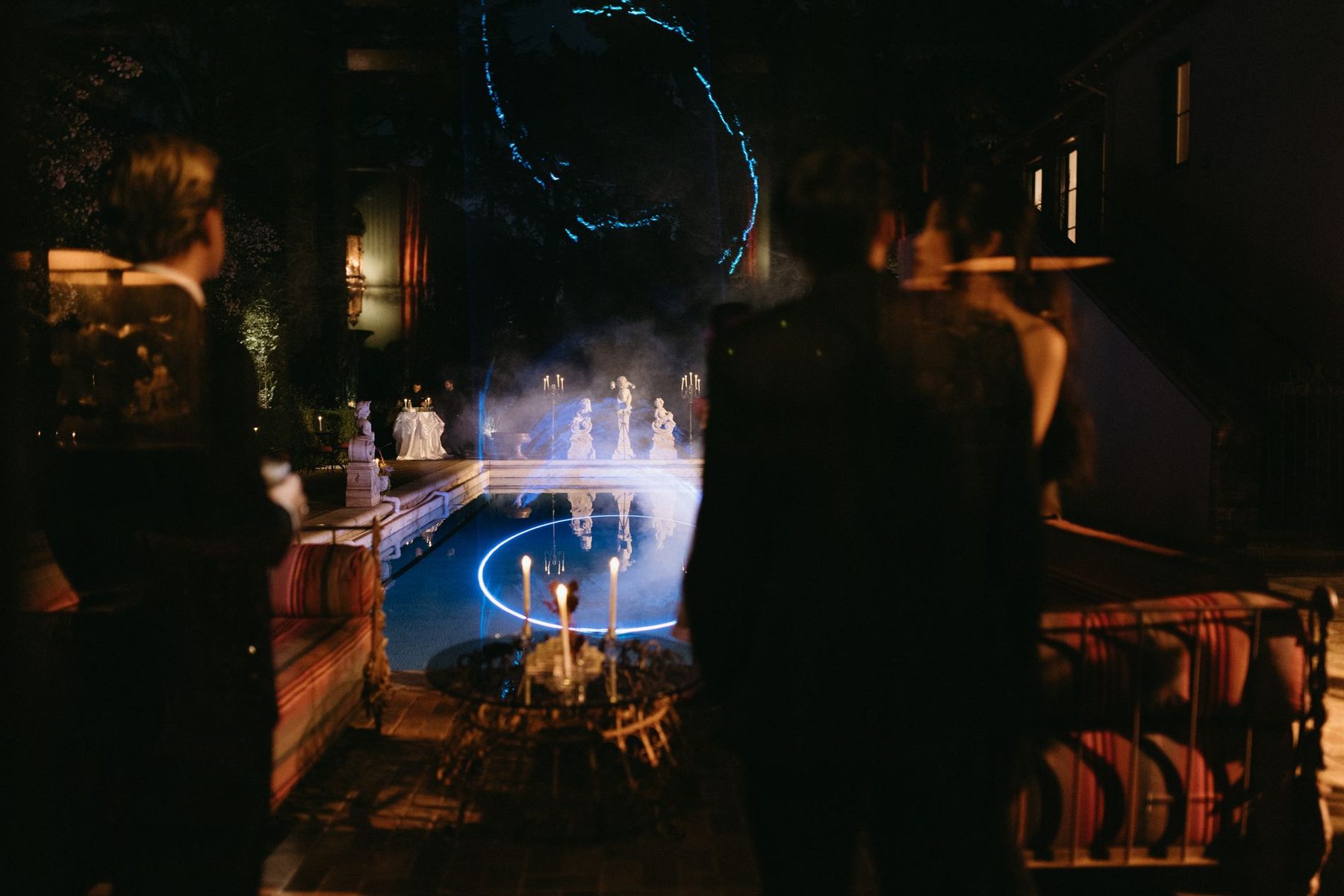 Guests around a pool ringed with blue light beneath candlelit statuary